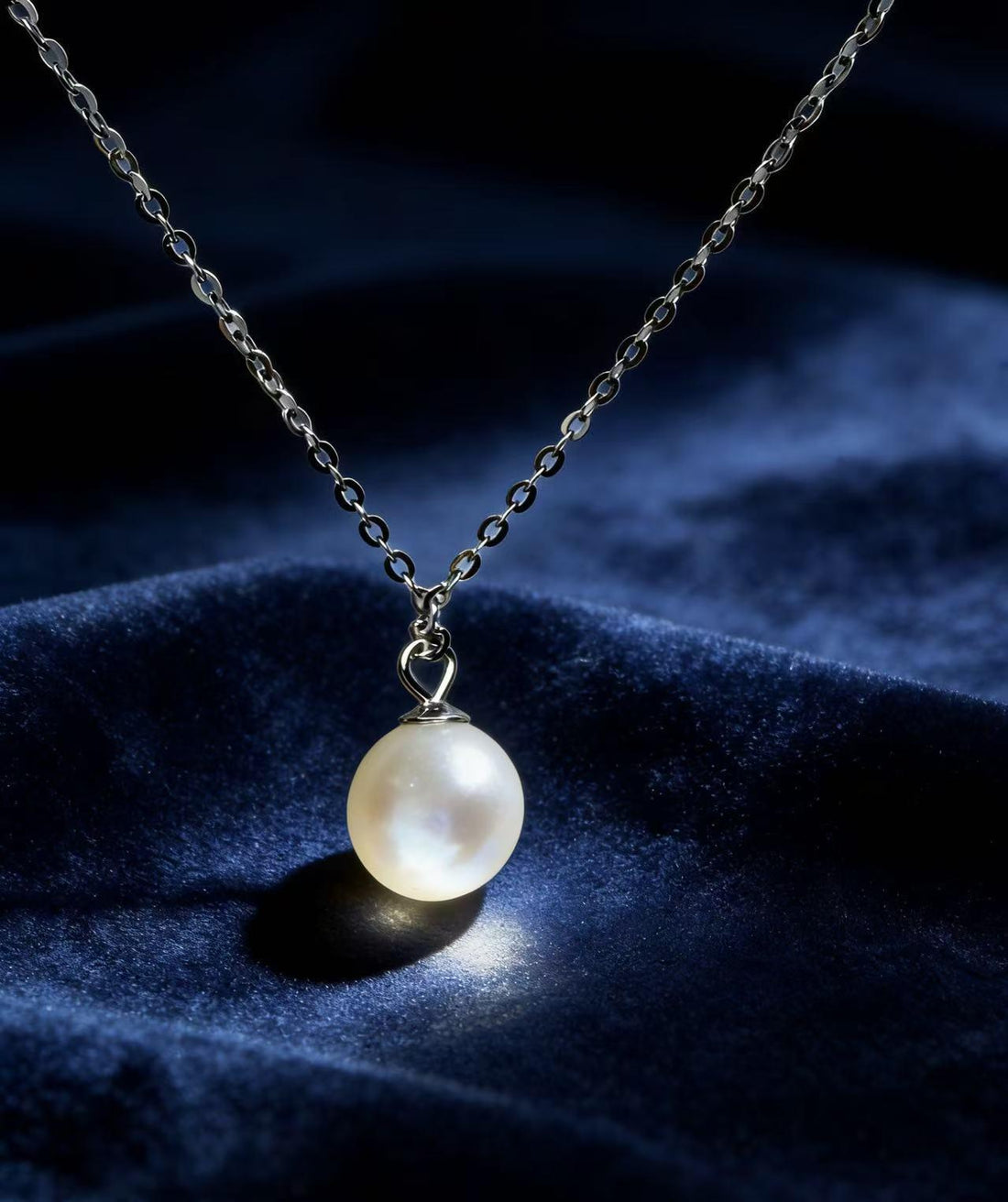 A single large round white pearl pendant on a sterling silver chain, displayed on blue velvet fabric under a spotlight.