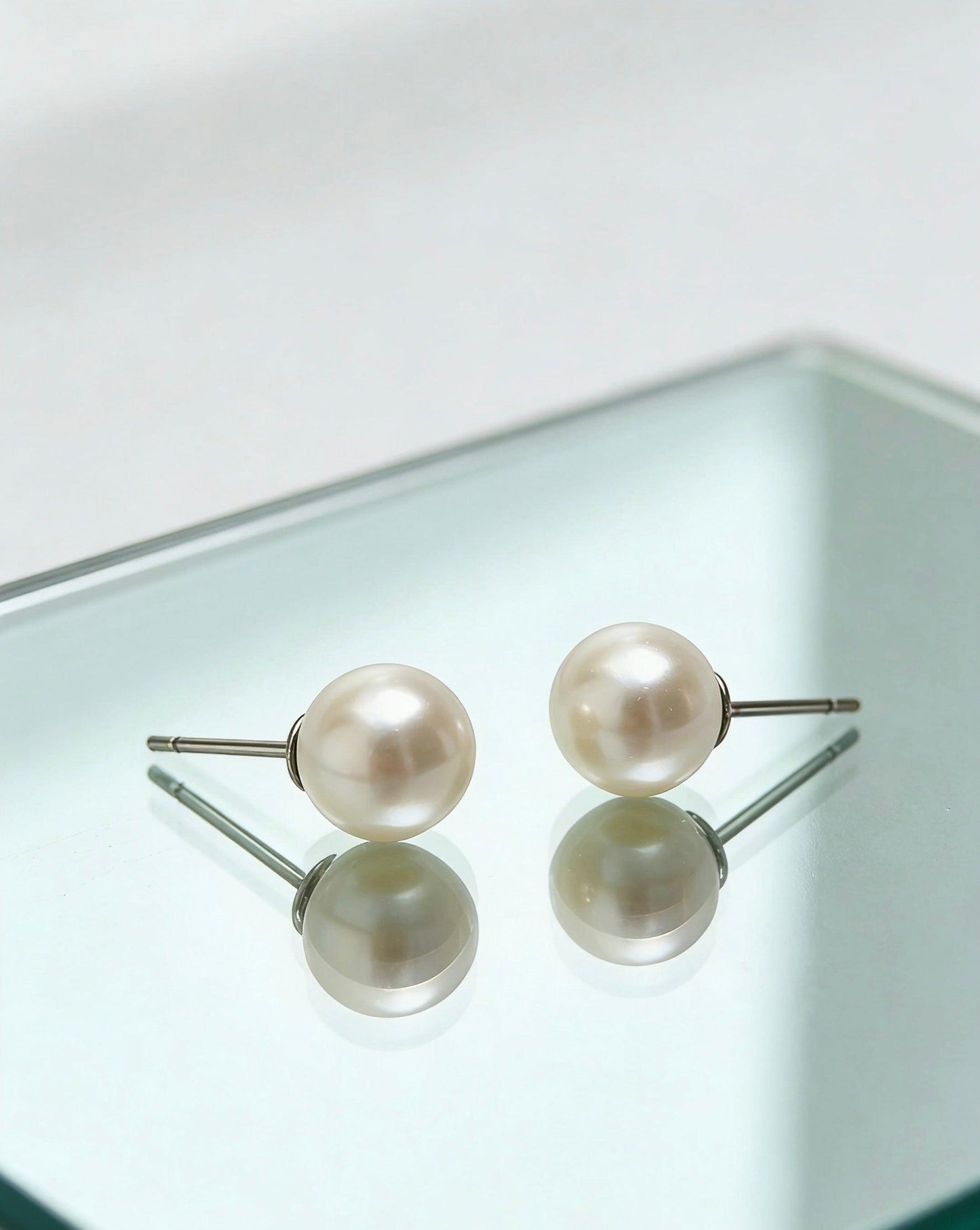 Two 7mm natural freshwater pearl stud earrings with sterling silver posts, reflecting on a glass surface.