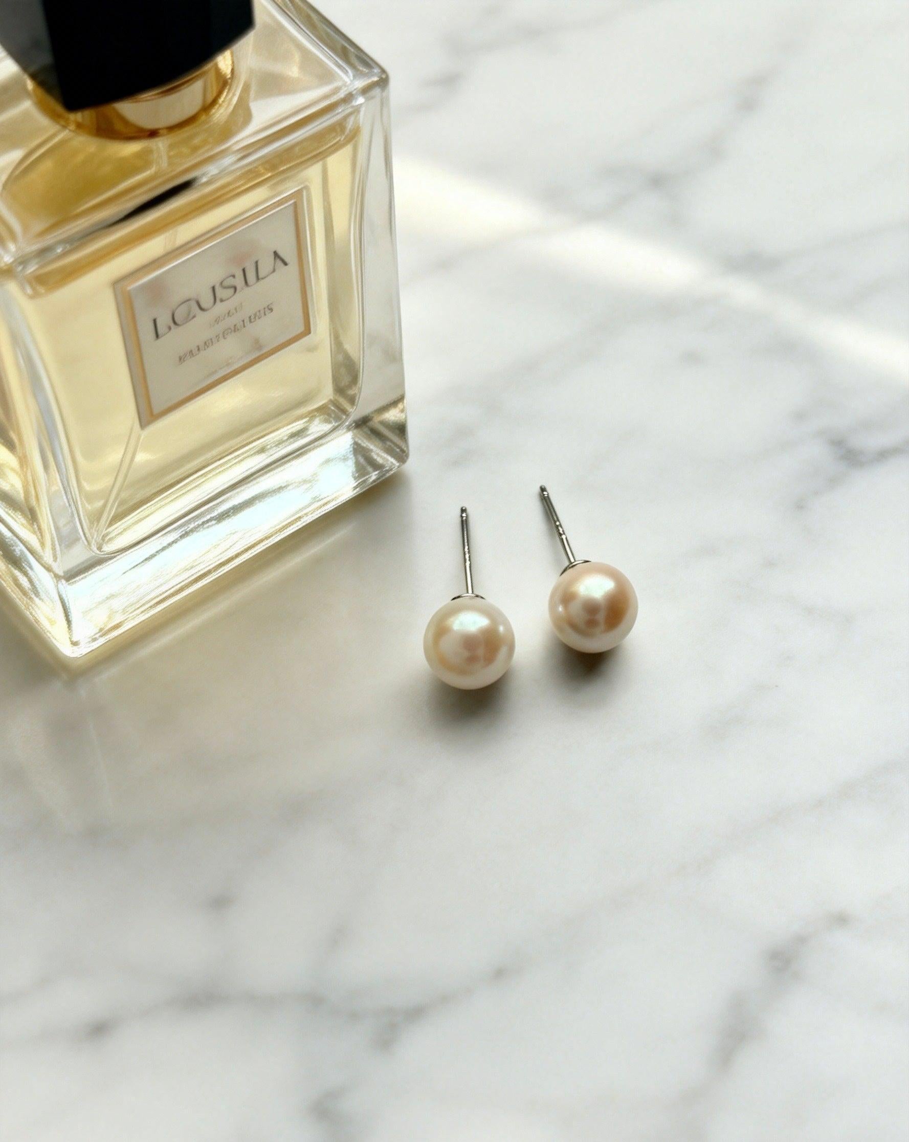 Two Luvymia 7mm natural saltwater Hepu pearl stud earrings with sterling silver posts on marble next to perfume.