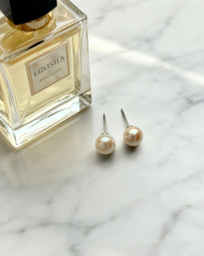 Two Luvymia 7mm natural saltwater Hepu pearl stud earrings with sterling silver posts on marble next to perfume.